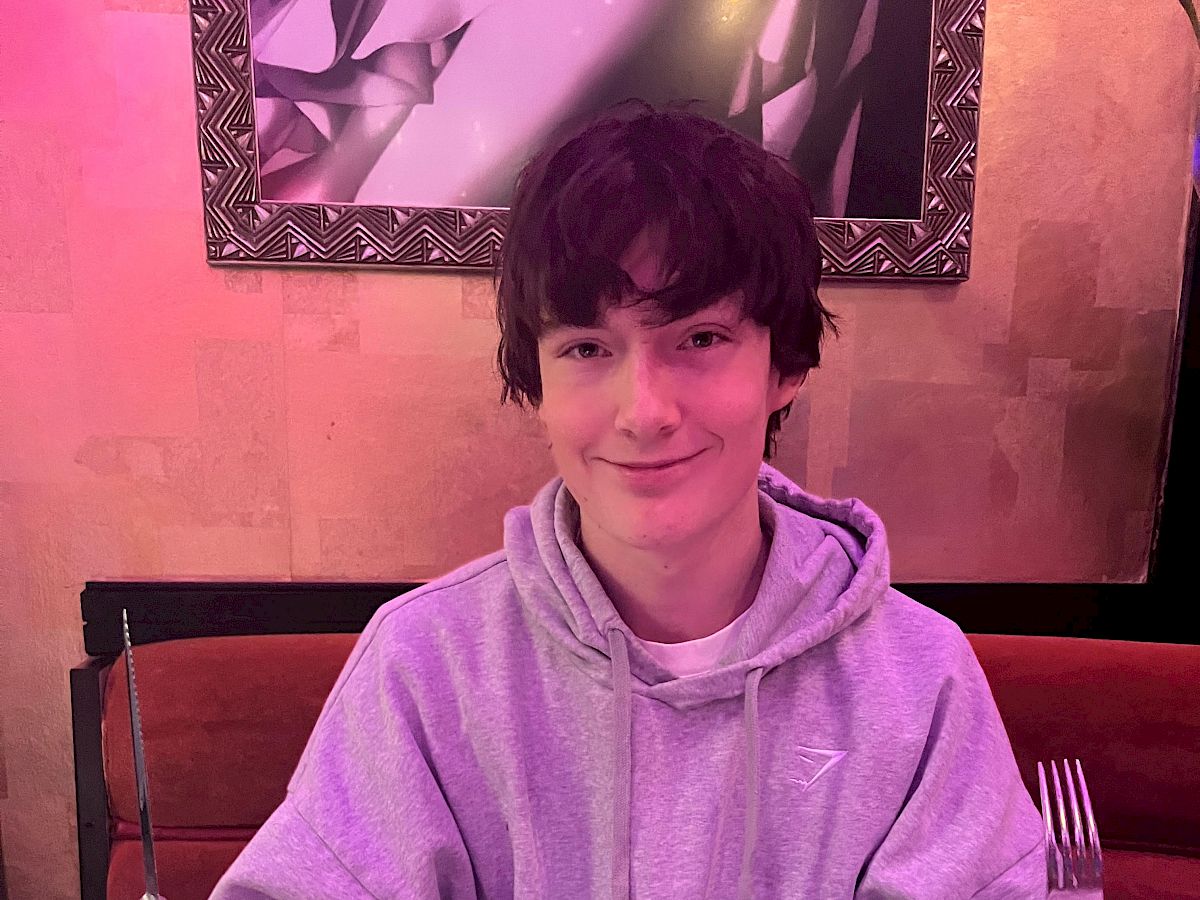 A person in a hoodie sits at a table with a large steak and sides, holding utensils, ready to eat.