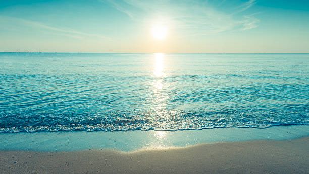 A serene beach scene at sunset with calm turquoise waves, glowing sun low on the horizon, gentle surf kissing the sandy shore, and clear sky.