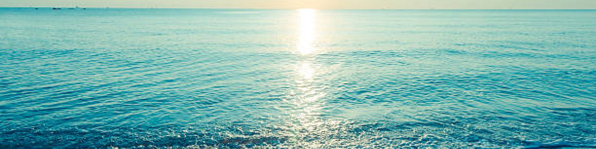 A tranquil beach scene with gentle waves, a clear sky, and the sun shining over the horizon, casting reflections on the water.