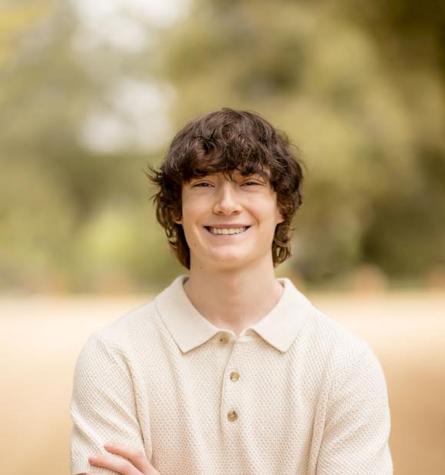 A person with curly hair and a beige shirt crosses their arms and smiles in an outdoor setting.