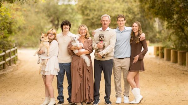 A family of eight stands on a dirt path in a park, smiling and posing with two small dogs, all dressed in light, casual outfits.