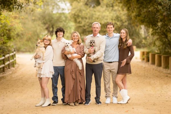 A family of five, with each holding a small dog, stands on a tree-lined path, dressed in casual and warm attire, sharing smiles and affection.
