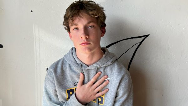 A young man sits on a wooden bench against a wall with cursive writing, wearing a gray hoodie and beige pants, hand over his chest.